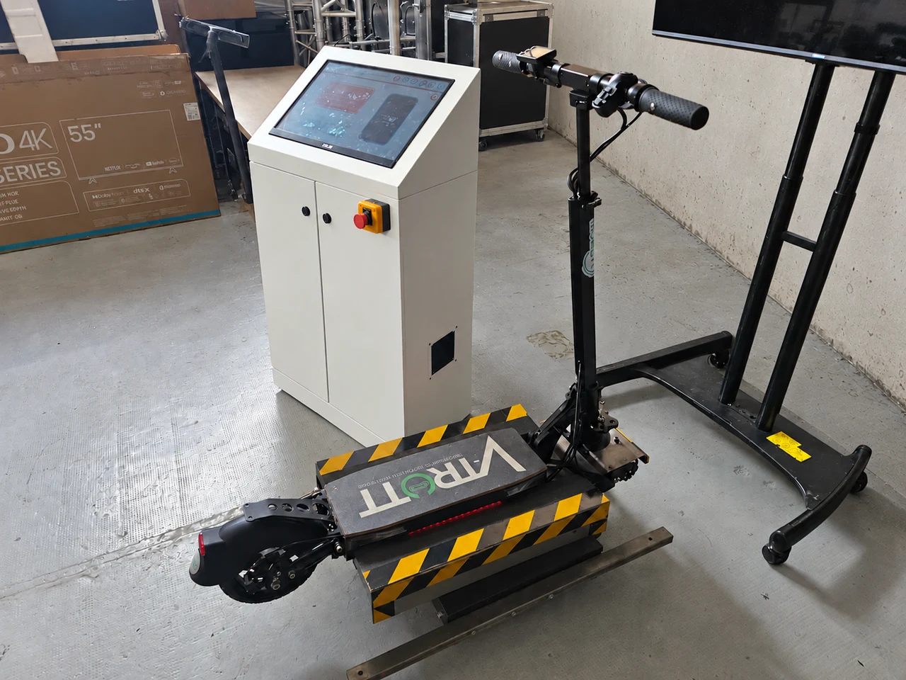 Trottinette VTrott sur une plateforme noire à bandes jaunes, avec borne tactile blanche et écran sur pied